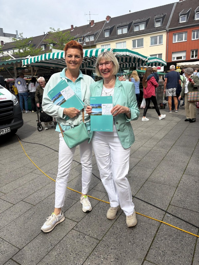 Ute Schmidt und eine Leserin halten die Sommerausgabe 2026 der Kölner StadtteilLiebe auf dem Rodenkirchener Markt