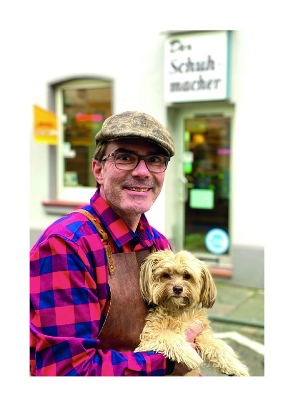 Andreas Wölfer vor seiner Werkstatt Der Schuhmacher in Köln-Rodenkirchen, mit Hund auf dem Arm