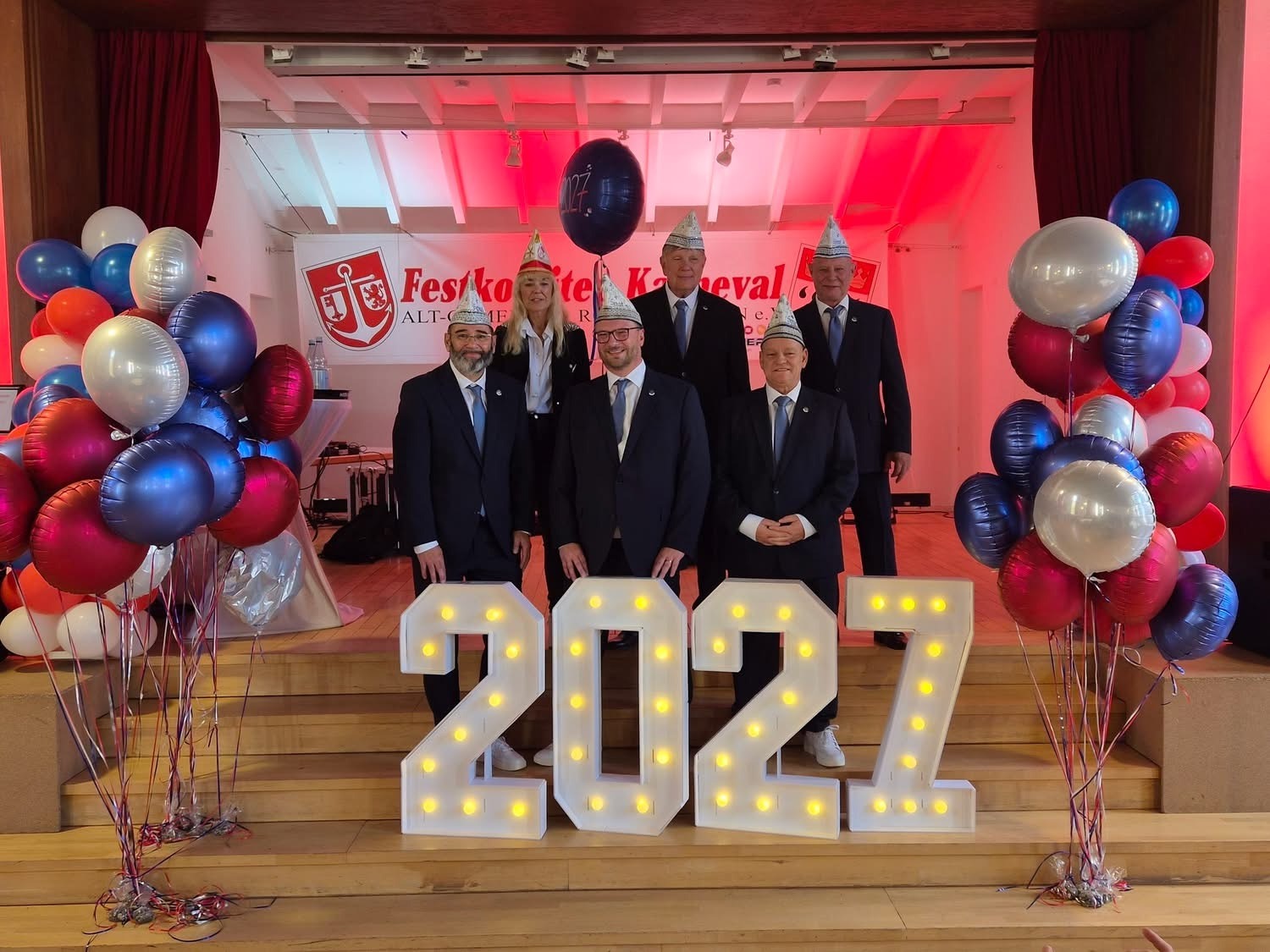 Designiertes Dreigestirn Rodenkirchen 2027 mit Begleitteam vor leuchtender Jahreszahl im Gemeindesaal.