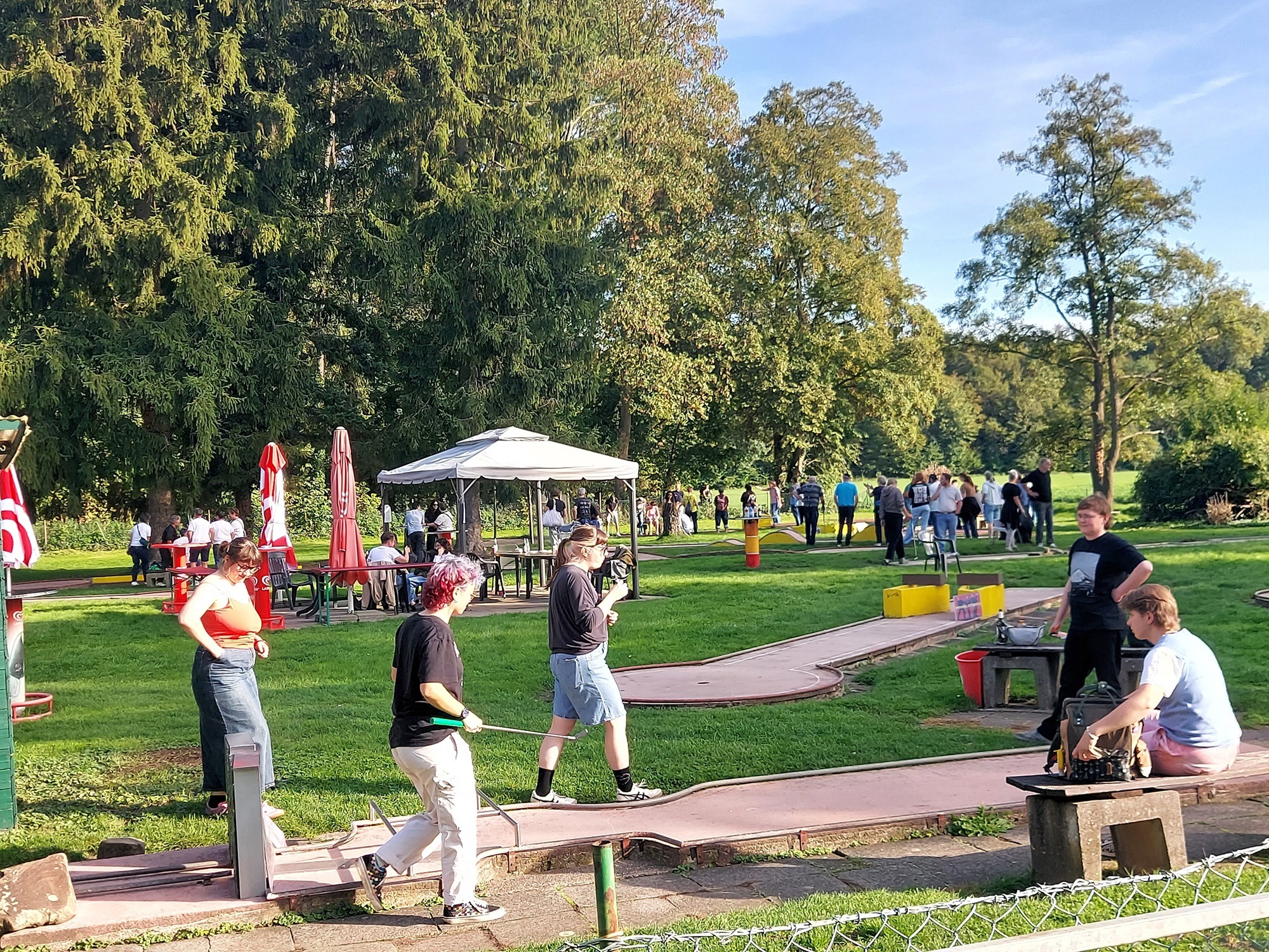 Gäste beim Queer-Minigolf-Turnier in Rodenkirchen zwischen Bahnen, Pavillon und Bäumen in der Sonne