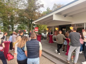 Gäste hören die Eröffnungsansprache vor dem Vereinsziegel von Rot-Weiss Zollstock